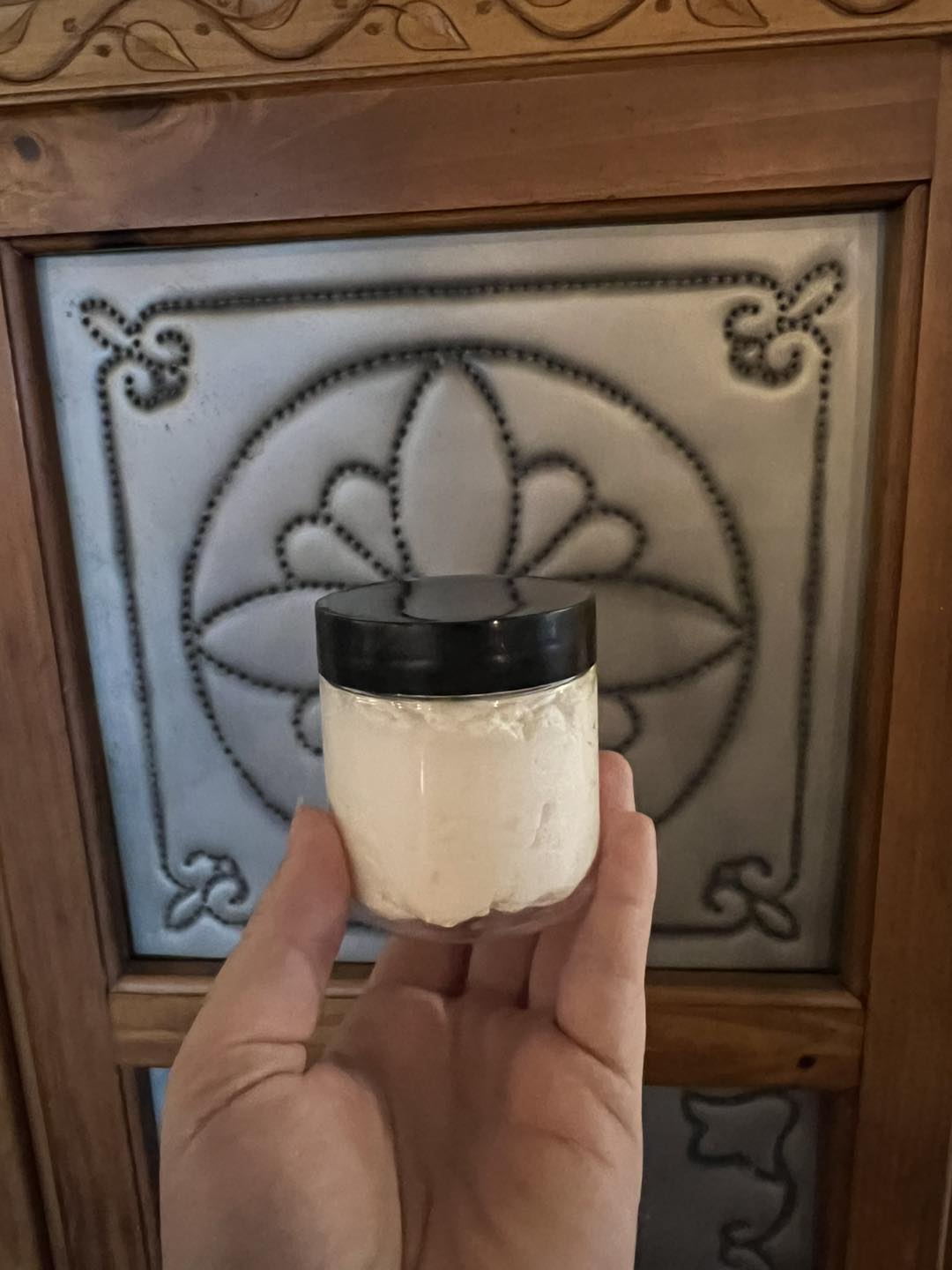 Hand holding a small jar of cream with a black lid in front of a decorative wooden panel, highlighting the product's texture and packaging in a cozy setting.