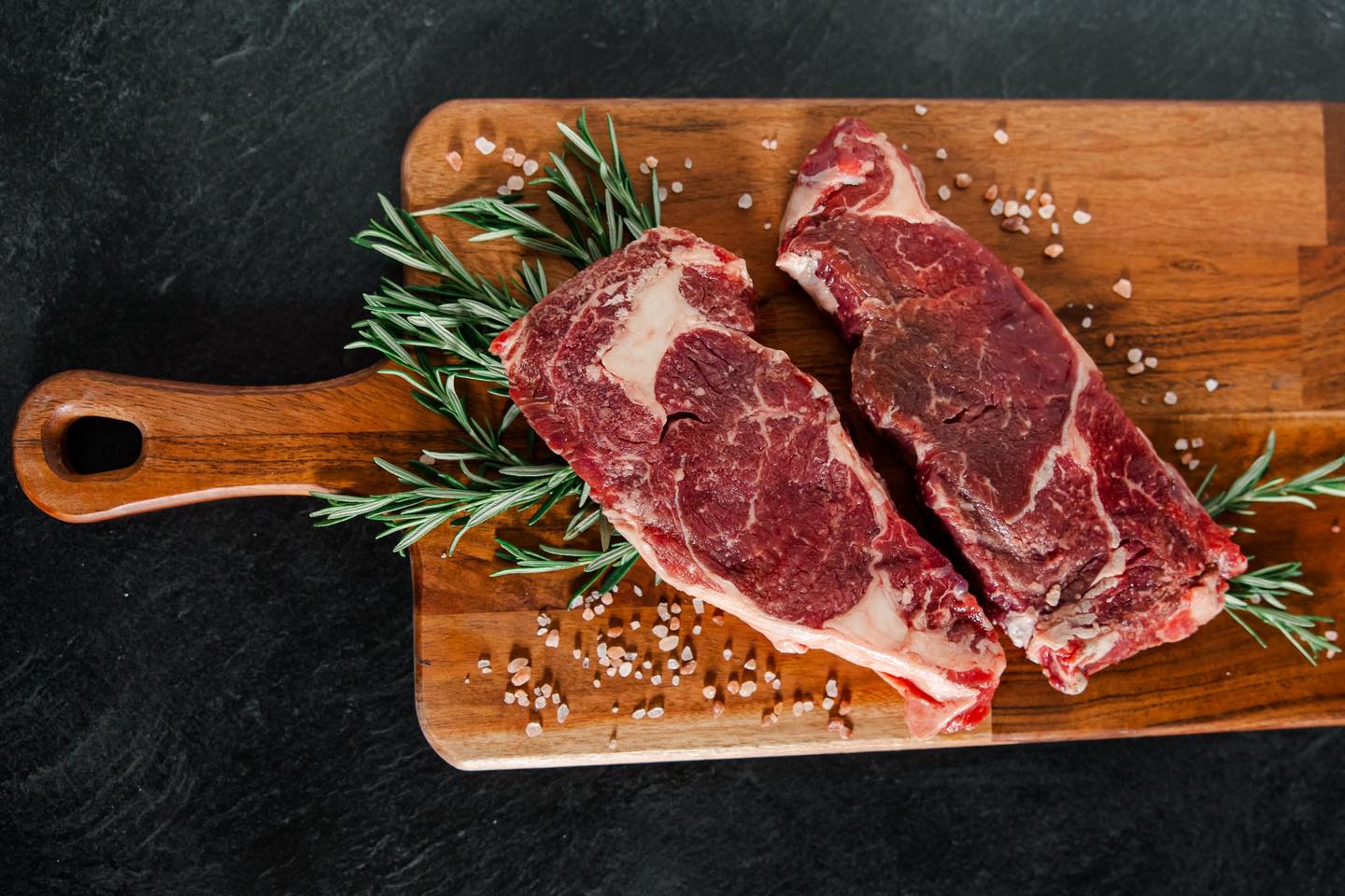 Raw marbled beef steaks resting on a wooden cutting board, garnished with fresh rosemary and sprinkled with coarse sea salt, set against a dark background.
