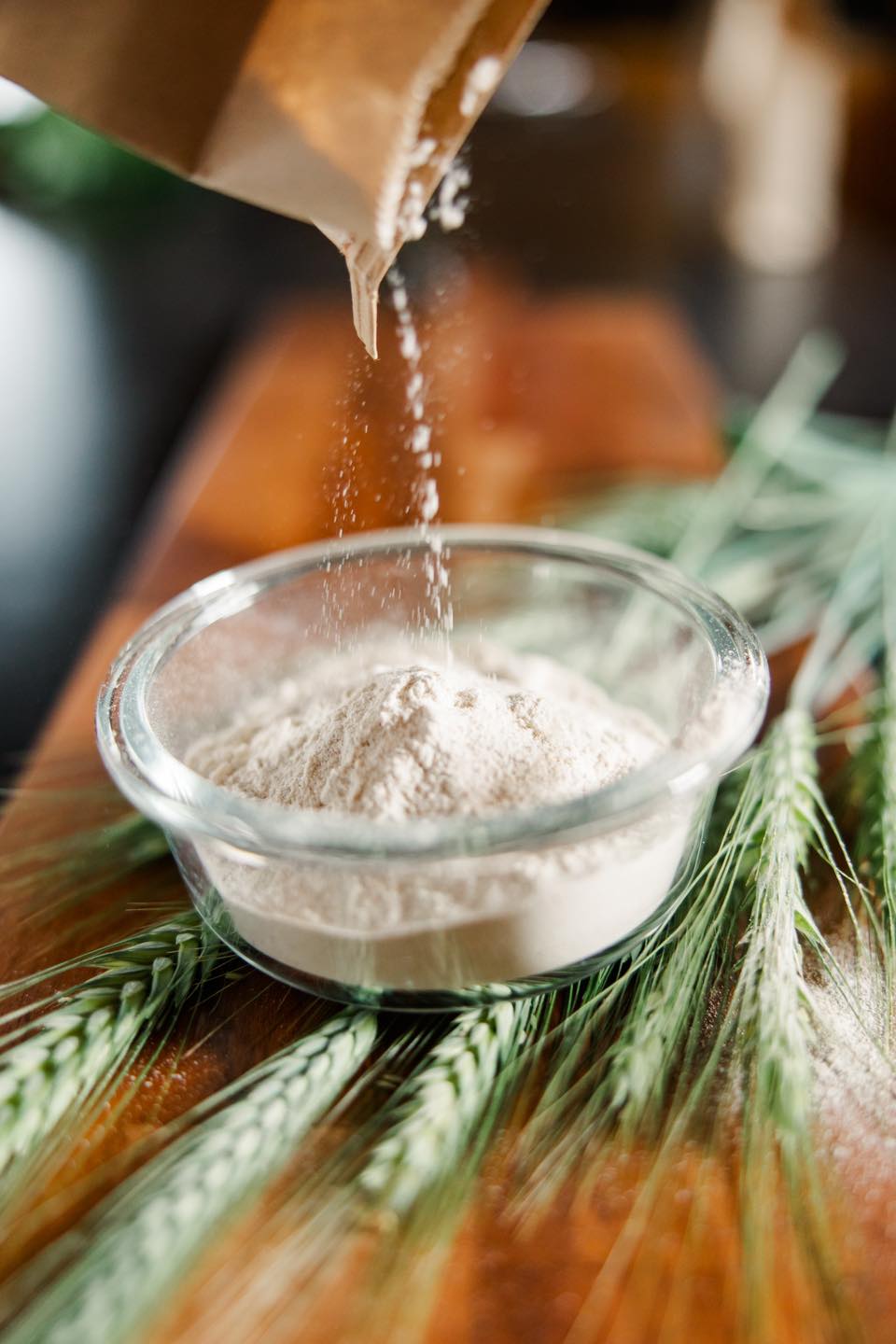 A close-up image of whole wheat flour being poured from a paper bag into a glass bowl, surrounded by fresh wheat stalks on a wooden surface, highlighting the natural ingredients used in baking and cooking.