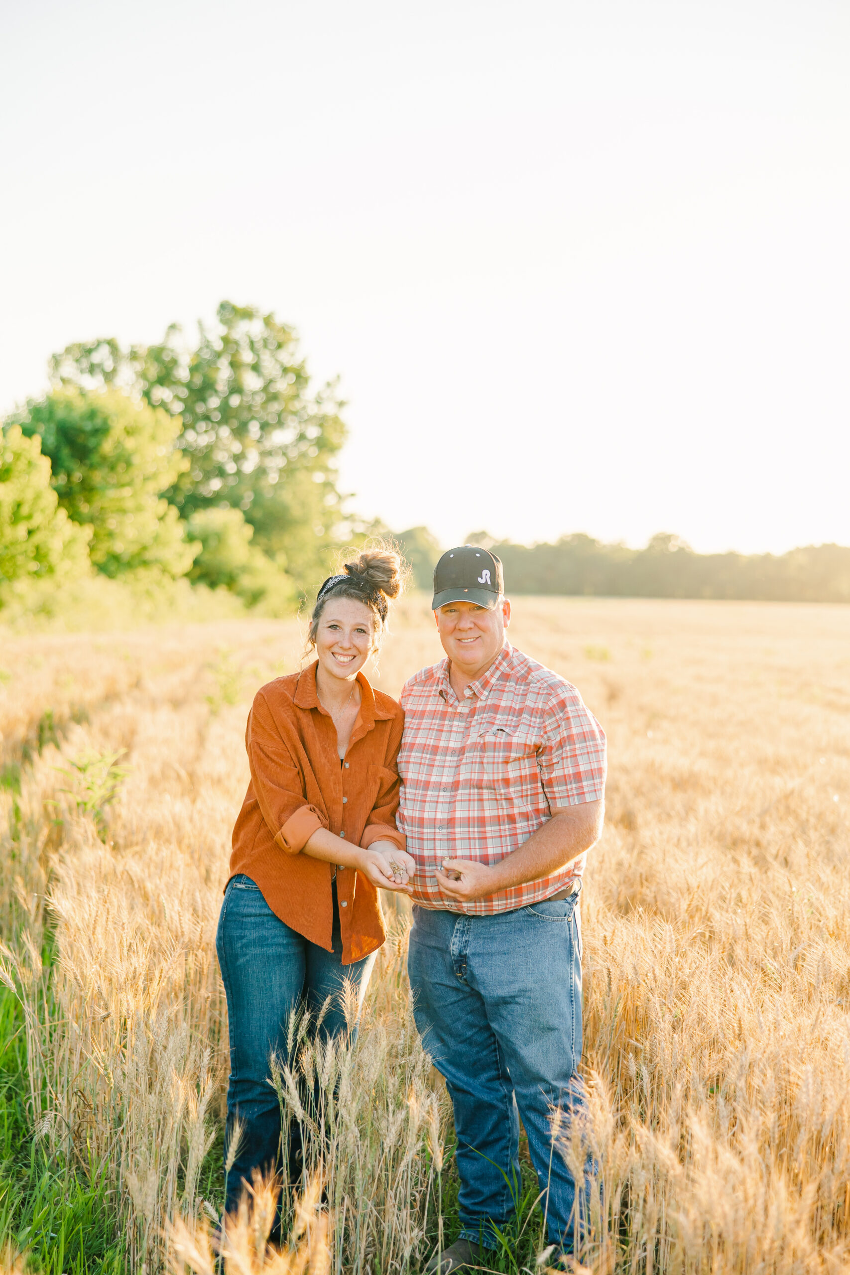 A smiling couple stands in a golden wheat field during sunset, holding grains in their hands. The woman wears a rust-colored shirt and blue jeans, while the man is dressed in a plaid shirt and a cap. Lush greenery and a clear sky enhance the scenic agricultural backdrop.