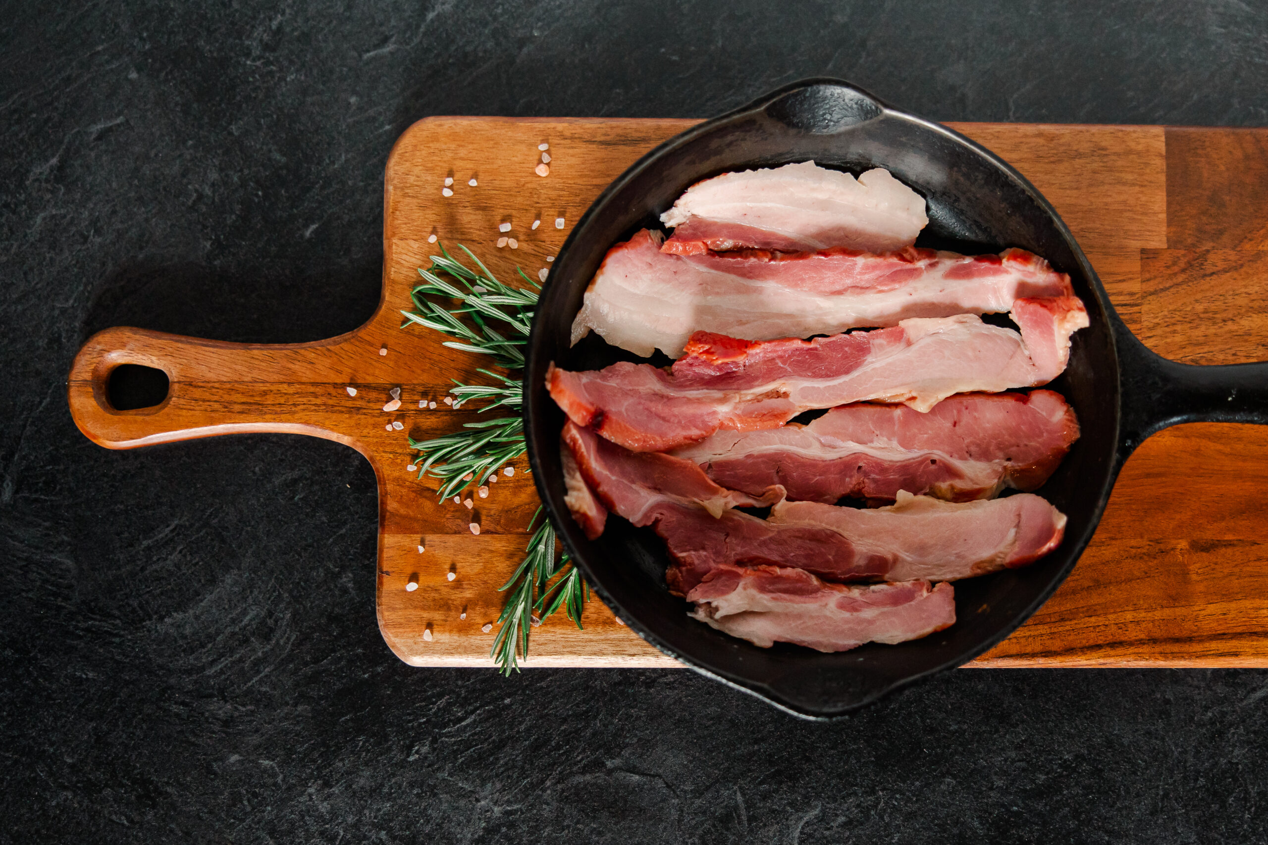 Sliced bacon in a cast iron skillet resting on a wooden cutting board, garnished with fresh rosemary and pink salt crystals.