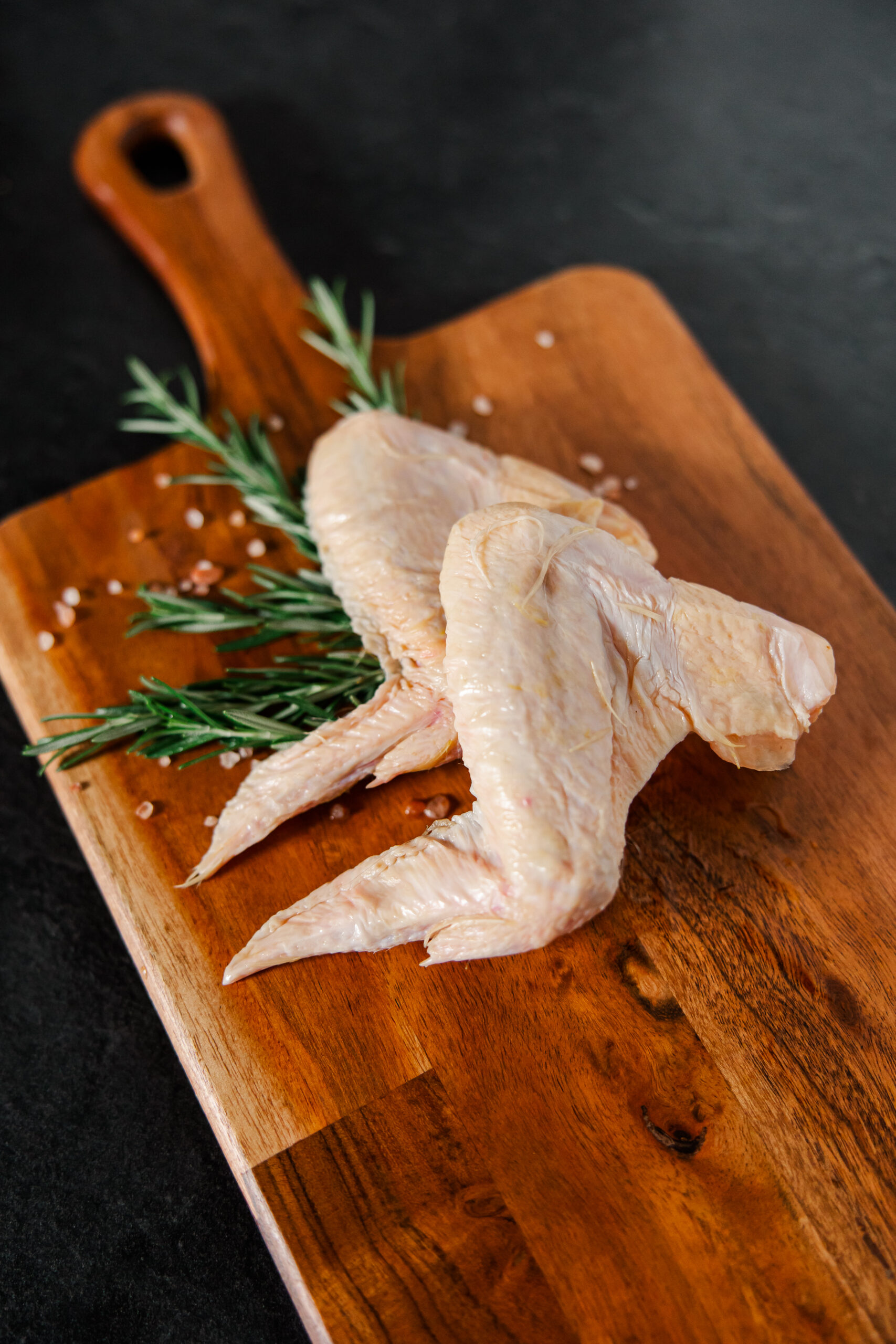 Raw chicken wings placed on a wooden cutting board with fresh rosemary and pink salt, ideal for meal preparation and cooking inspiration.