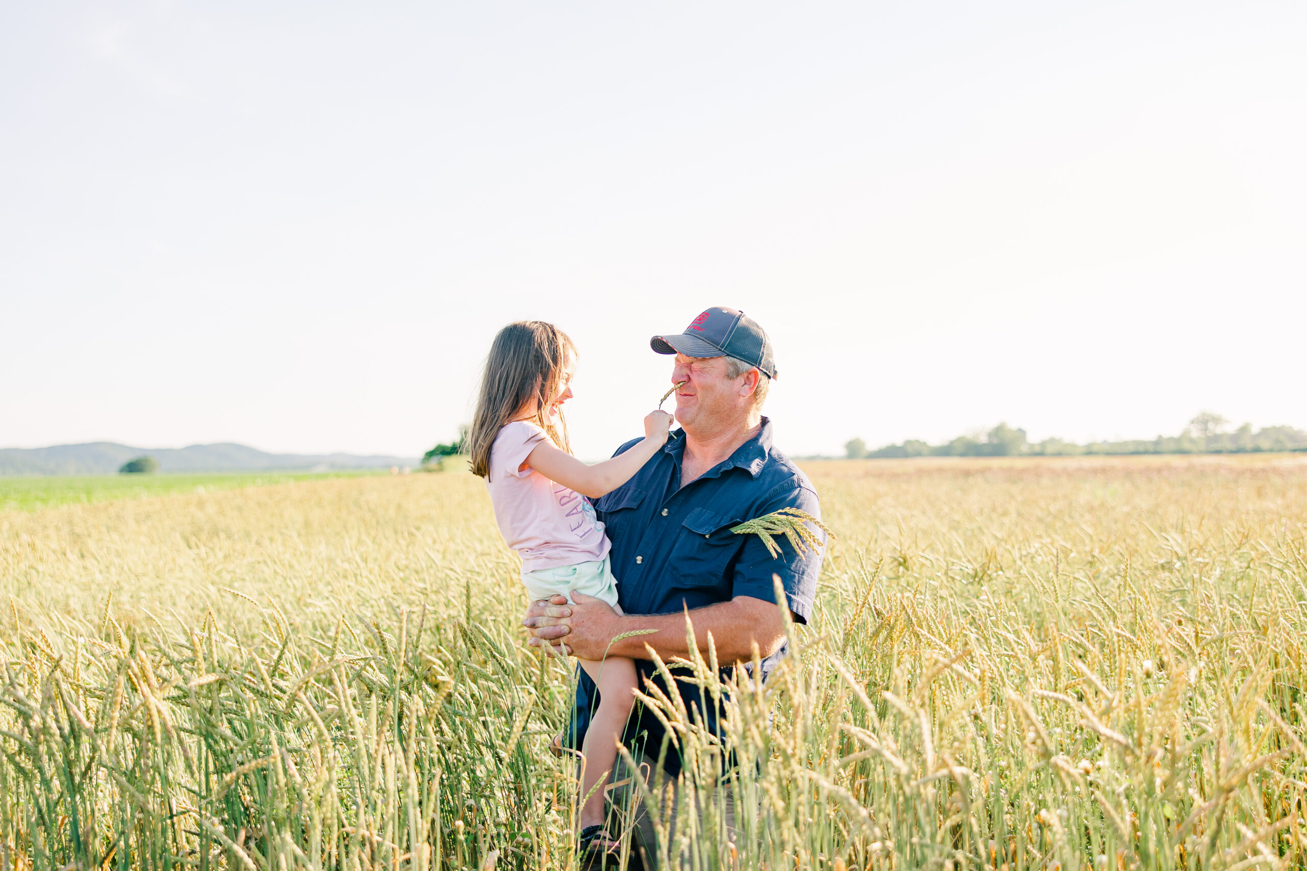 A farmer holds a young girl in a wheat field, both smiling as they share a playful moment under a clear blue sky. The tall wheat sways gently in the breeze, showcasing a vibrant rural landscape.